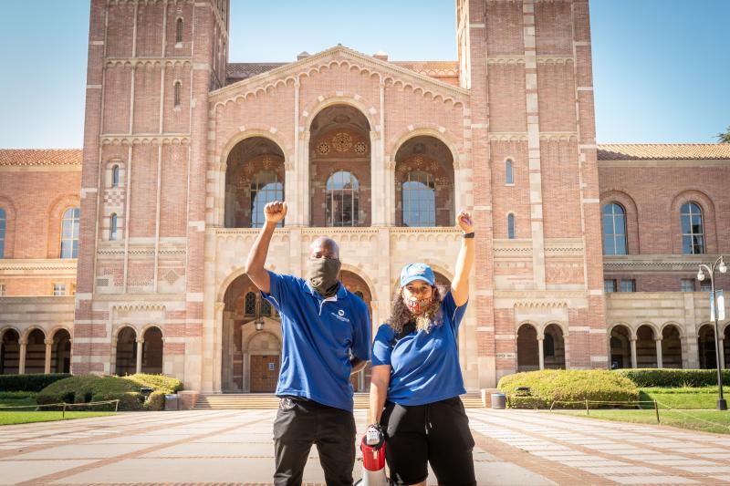 Black male and female UCLA staff wearing blue polos standing side by side in front of Royce Hall with opposing fists raised