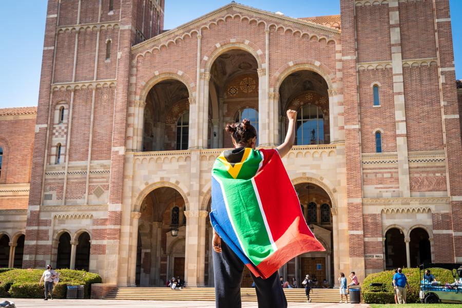Female student donning South African flag as a cape and raising her right arm in a fist in front of Royce Hall at UCLA